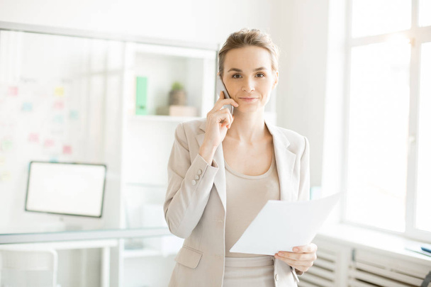 Giovane elegante dipendente con smartphone e carta che parla con i clienti durante la giornata lavorativa in ufficio
 - Foto, immagini