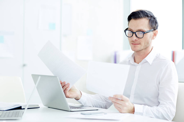Young businessman in eyeglasses and white shirt looking through financial documents by workplace - Foto, imagen