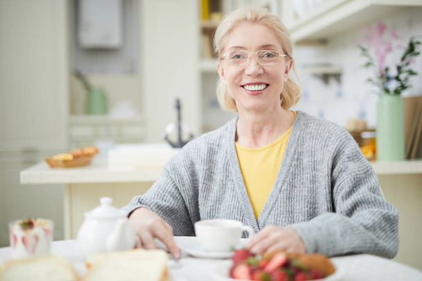 Feliz mujer mayor casual mirando a la cámara con sonrisa dentada mientras toma el té en casa
 - Foto, Imagen