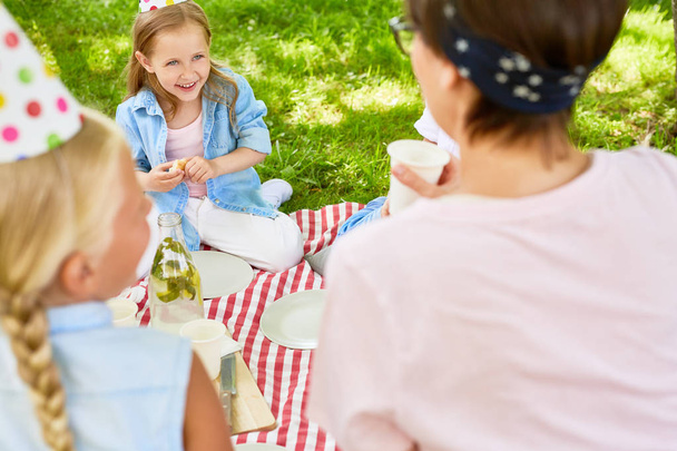 Cheerful little girl with piece of sandwich talking to her teacher and friend during picnic in summer park - Photo, Image