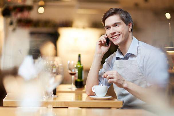 Joven camarero hablando por teléfono durante su descanso para tomar café en el trabajo
 - Foto, Imagen