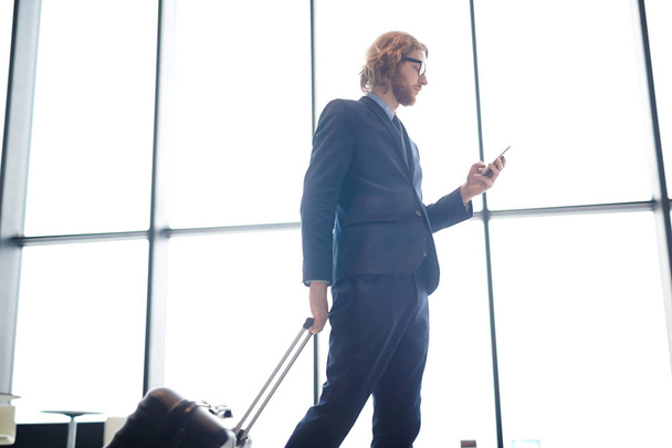 Νεαρός άνδρας στο κομψό κοστούμι μηνυμάτων στο smartphone και το τράβηγμα βαλίτσα ενώ τα πόδια στο σιδηροδρομικό σταθμό πτήσης - Φωτογραφία, εικόνα