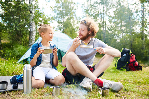 Jonge scout en zijn zoon of vriend reis eten zittend op het gras niet ver van hun tent - Foto, afbeelding