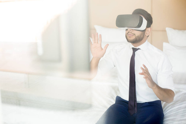 Сучасний бізнесмен в формальному одязі сидить на ліжку і дивиться конференцію або презентацію в гарнітурі віртуальної реальності
 - Фото, зображення
