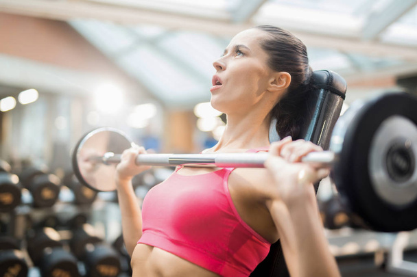 Jovem mulher tentando levantar pesado barbell de seu peito enquanto exercitando no ginásio
 - Foto, Imagem