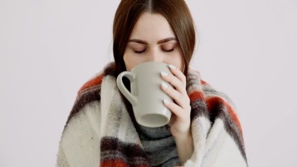a beautiful cold girl of Eurarean appearance in a woolen blanket is drinking hot tea by the white wall. - Footage, Video