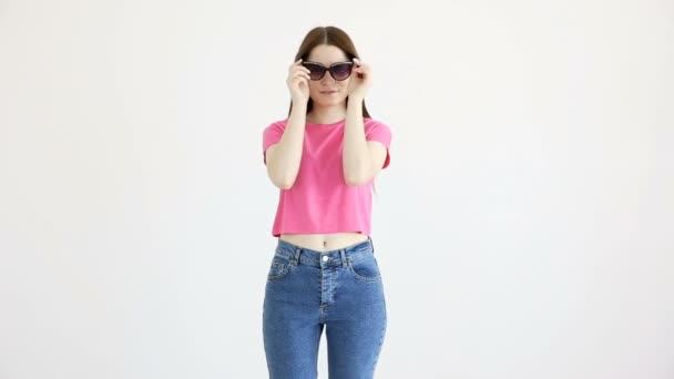 mooi vrolijk meisje in zonnebrillen, roze top en jeans poseren tegen de witte muur - Video