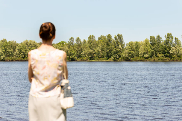 A woman with a chignon, standing up close to the Dnieper river in Kiev, Ukraine, observes the trees in the distance. The silhouette of the lady is out of focus, against a focused background. - Zdjęcie, obraz