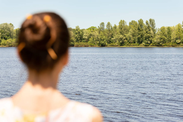 A woman with a chignon, standing up close to the Dnieper river in Kiev, Ukraine, observes the trees in the distance. The silhouette of the lady is out of focus, against a focused background. - Фото, изображение