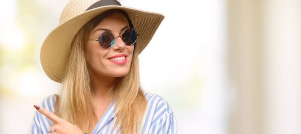 Nuori nainen yllään aurinkolasit ja kesän hattu osoittaa poispäin sormella
 - Valokuva, kuva