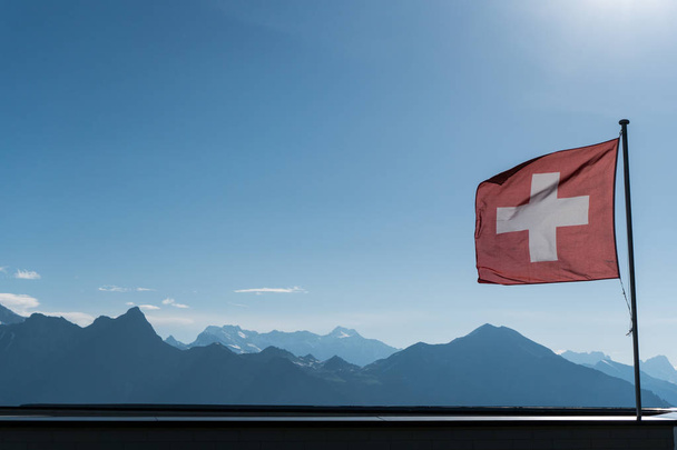 Szwajcarska flaga wiejący wiatr z gorgoues góry krajobraz i niebieski niebo za - Zdjęcie, obraz
