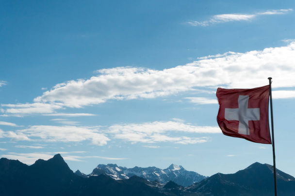 Szwajcarska flaga wiejący wiatr z gorgoues góry krajobraz i niebieski niebo za - Zdjęcie, obraz