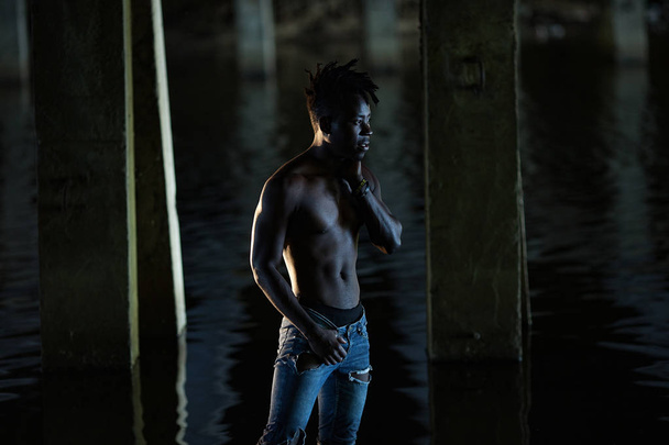 Afryki młody człowiek z bare klatce w dżinsy stoi w wodzie pod mostem na tle betonowych podpór. - Zdjęcie, obraz