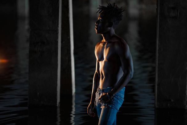 Young African man with bare-chested in jeans stands in water under bridge on background of concrete supports. - Zdjęcie, obraz