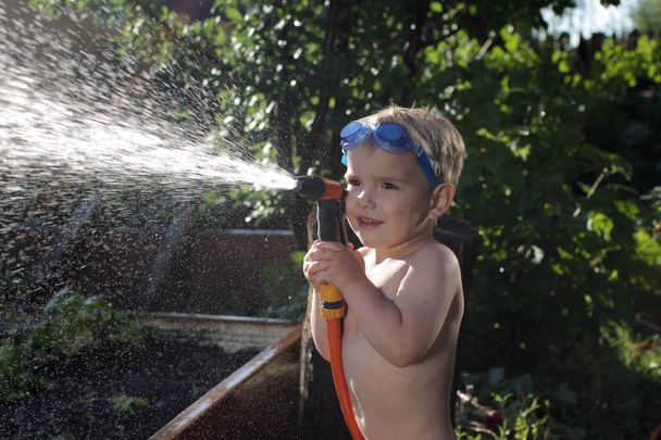 A happy little boy in the watersport goggles with a pistol in his hands watering the garden in the summer cottage during summer vacation, happy family summertime concept, outdoor lifestyle - Photo, Image
