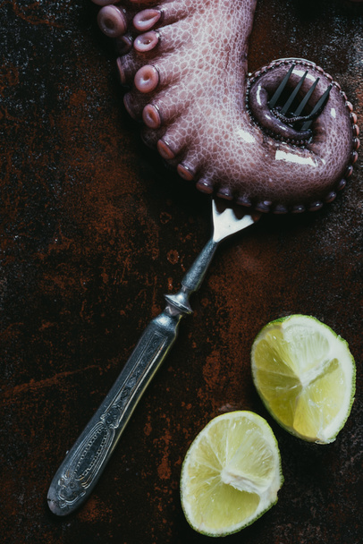 top view of big octopus tentacle with fork and limes on rusty metal surface - Foto, Bild