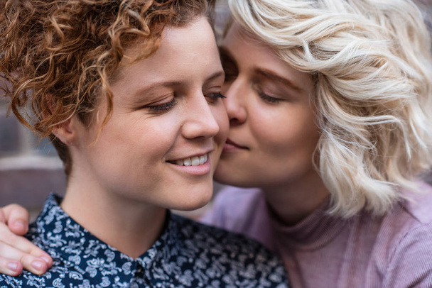 Closeup of a young woman affectionately kissing her smiling girlfriend on the cheek while standing together in front of a brick wall outside - Photo, image
