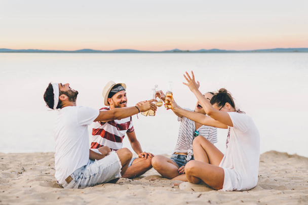 Group of happy young people sitting together at the beach talking and drinking beers - Foto, immagini