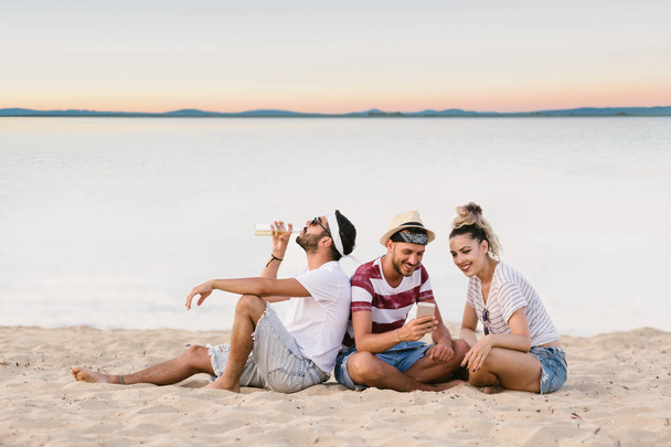 Group of happy young people sitting together at the beach and taking selfie - Foto, afbeelding