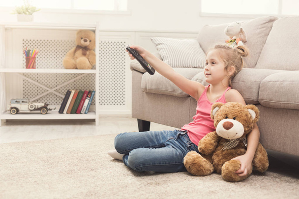 Little casual girl watching tv at home. Female kid sitting on floor carpet with her toy friend teddy bear, holding remote and switching channels, copy space - Photo, Image