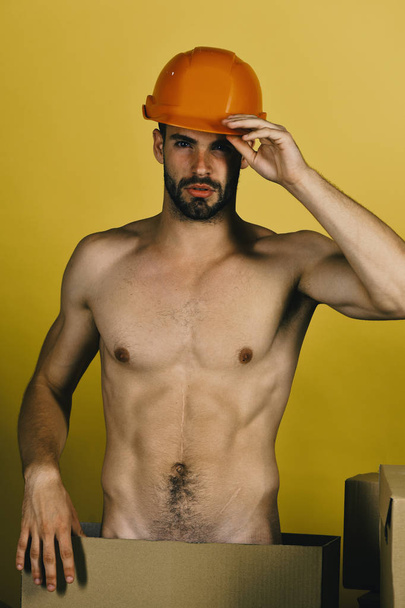 Man standing naked among cardboard boxes. Guy with sexy torso in hard hat on yellow background - Фото, изображение