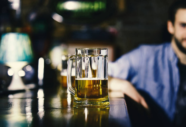 Glass of beer on wooden bar counter. Man sits behind glass of light beer. - Photo, Image