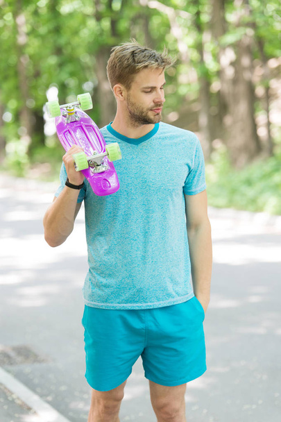 Still childish. Man calm face carries penny board park nature background, defocused. Guy carries penny board shoulder, ready to ride. Man likes to ride skateboard and sporty lifestyle. Hobby concept - Photo, Image