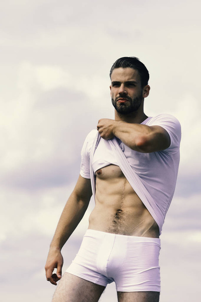 Man with unshaved and serious face on clear sky background. Macho wearing white tshirt and underwear demonstrates abs - Fotografie, Obrázek