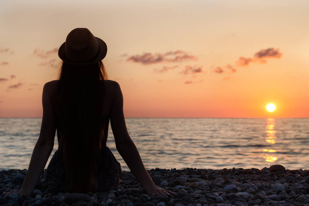 Mädchen mit Hut am Strand sitzend. Sonnenuntergang. Silhouette. zurück - Foto, Bild