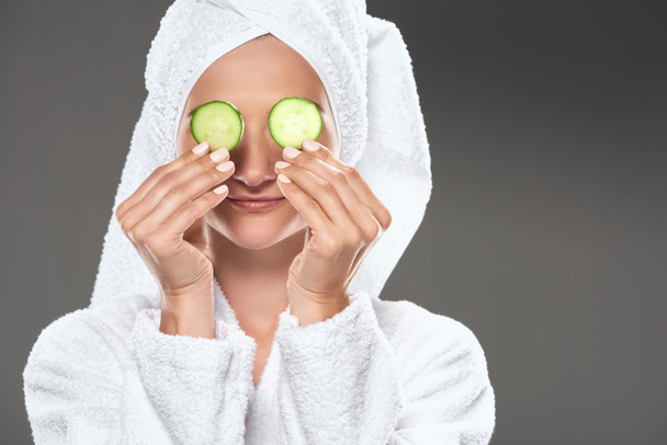 girl with clean skin and cucumber slices in white bathrobe and towel on head, isolated on grey - Foto, Bild