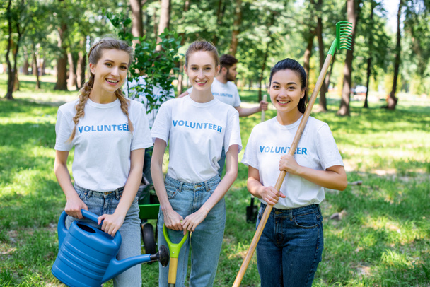 young female volunteers with watering can, shovel and rake standing in park  - Photo, Image