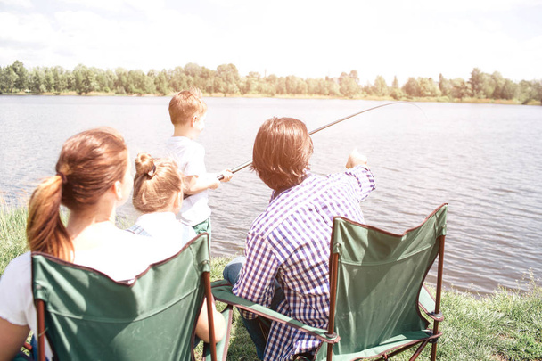A picture from the back. Small boy is holding a fish-rod and trying to catch some fish in the river. His dad is guiding him and pointing forward. Woman is holding small girl on her lap. - Photo, Image