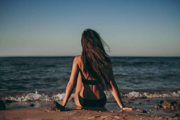 Девушка с ветром в волосах сидит на пляже и смотрит в горизонт
 - Фото, изображение