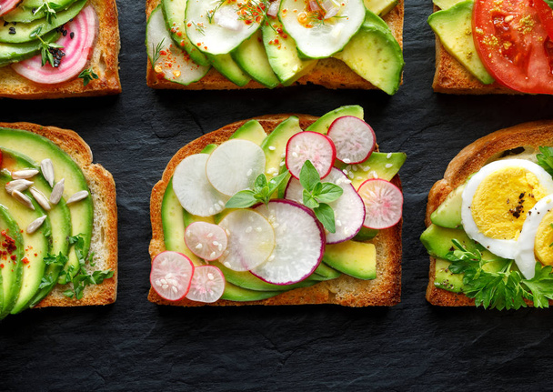 Avocado sandwiches, toasts with various vegetarian toppings on a black stone background, top view - Photo, Image