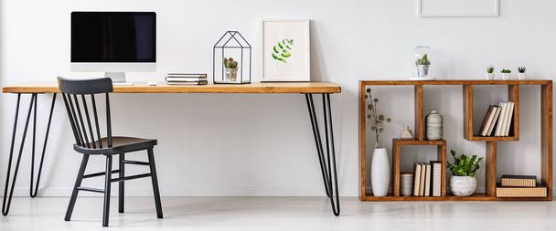 Hairpin table with computer, notebooks, poster and cactus standing in white home office interior with books and plants on wooden shelves - Fotó, kép