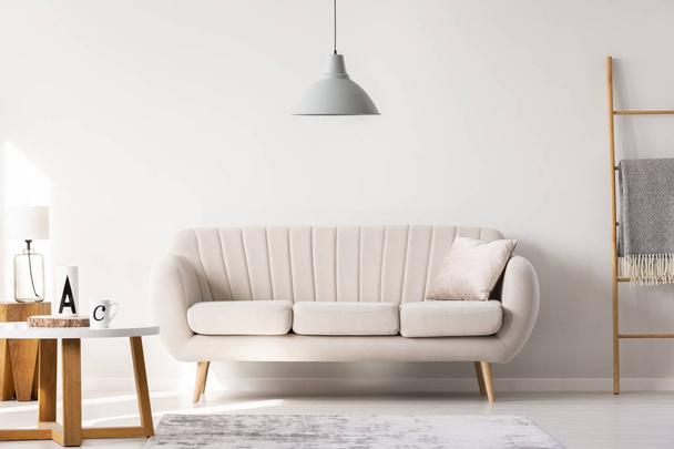 Real photo of a beige sofa standing in a simple living room interior under a lamp and next to a table and ladder - Photo, image