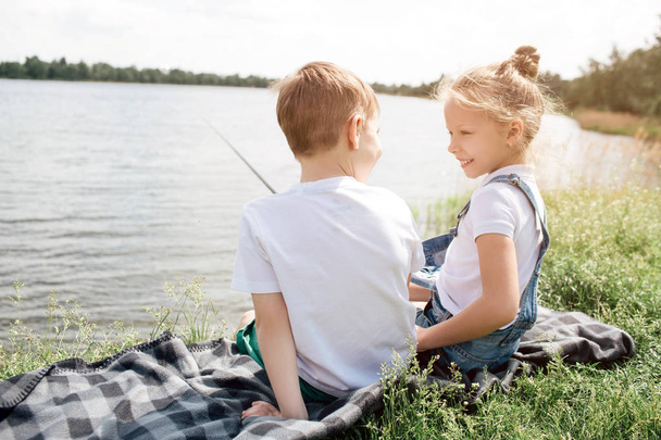 Une photo d'enfants assis ensemble sur une couverture et se regardant. La fille sourit. Elle tient une canne à poisson dans ses mains. Ils sont heureux.
. - Photo, image