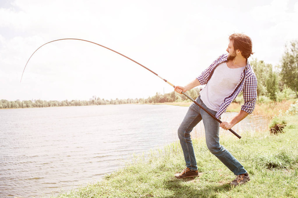 Calm and thoughtful guy is standing at the edge of lake and looking at it. He is holding fish-rod on his right shoulder with right hand. There is a beautiful weather outisde - Foto, Imagem