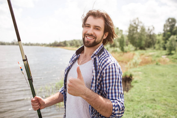 Calm and thoughtful guy is standing at the edge of lake and looking at it. He is holding fish-rod on his right shoulder with right hand. There is a beautiful weather outisde. - Foto, imagen