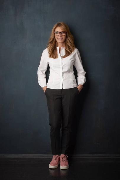 Full length shot of attractive mature businesswoman standing against at dark background while looking at camera and smiling.  - Fotografie, Obrázek