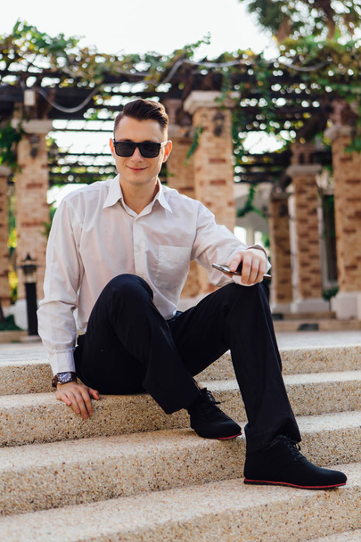 Young businessman holding his phone and sitting on stairs outdoors in a tropical country - 写真・画像