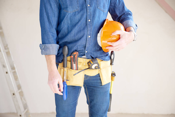 Cropped shot of a cunstruction man with handyman belt standing at construction site while holding safety helmet in his hand. - Fotografie, Obrázek