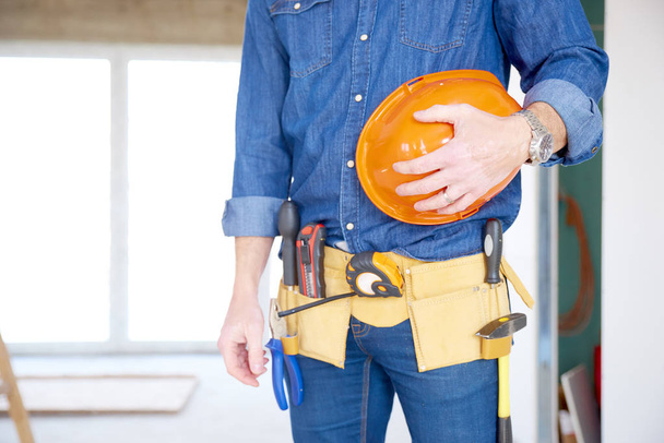 Cropped shot of a cunstruction man with handyman belt standing at construction site while holding safety helmet in his hand. - Photo, Image