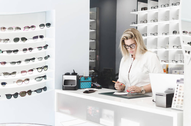Charismatic, confident oculist wearing white uniform. Woman wearing fashionable lenses. Doctor is standing in front of transparent stand with stylish, modern optical eyeglasses. - Photo, image