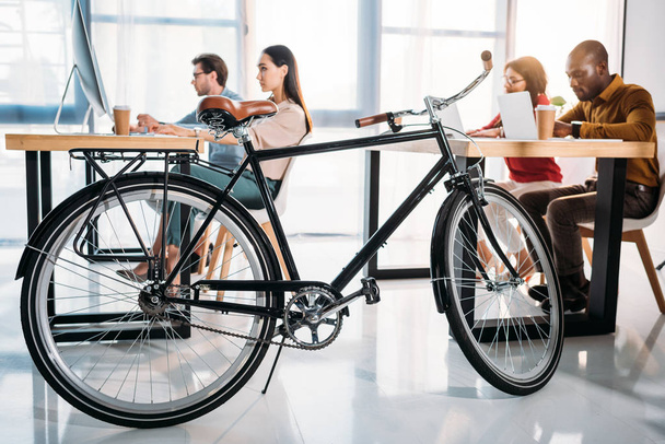 вид сбоку на велосипед и мультикультурных бизнесменов, работающих в офисе
 - Фото, изображение