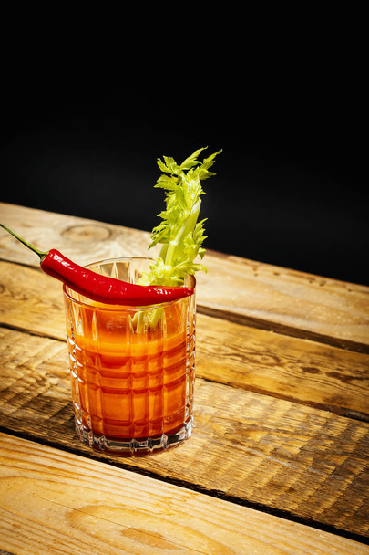 juicy red drink bloody mary with a piece of green celery and chilli peppers on a wooden table - Fotó, kép