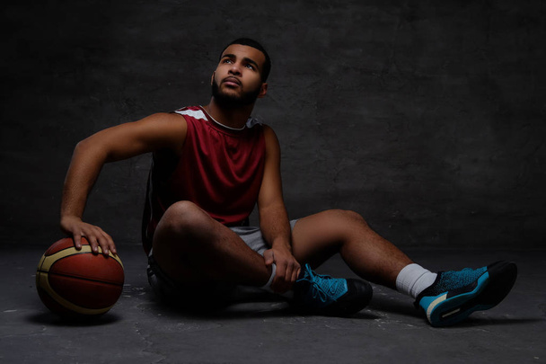 ボールを床に座ってスポーツウェアの若いアフリカ系アメリカ人のバスケット ボール選手。暗い背景上分離.  - 写真・画像