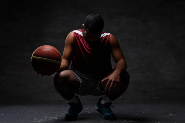 スポーツ ウエア、ボールとのしゃがむでアフリカ系アメリカ人のバスケット ボール選手。暗い背景上分離.  - 写真・画像