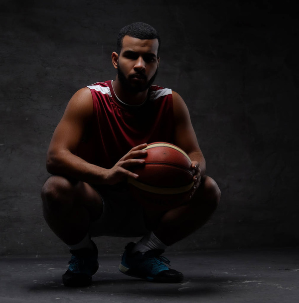 African-American basketball player in sportswear squatting with a ball. Isolated over dark background.  - Φωτογραφία, εικόνα