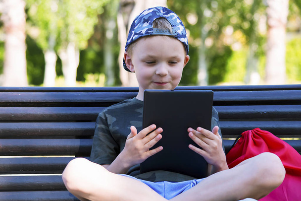 cute little boy is playing in the tablet on a bench in the summer - Foto, Bild